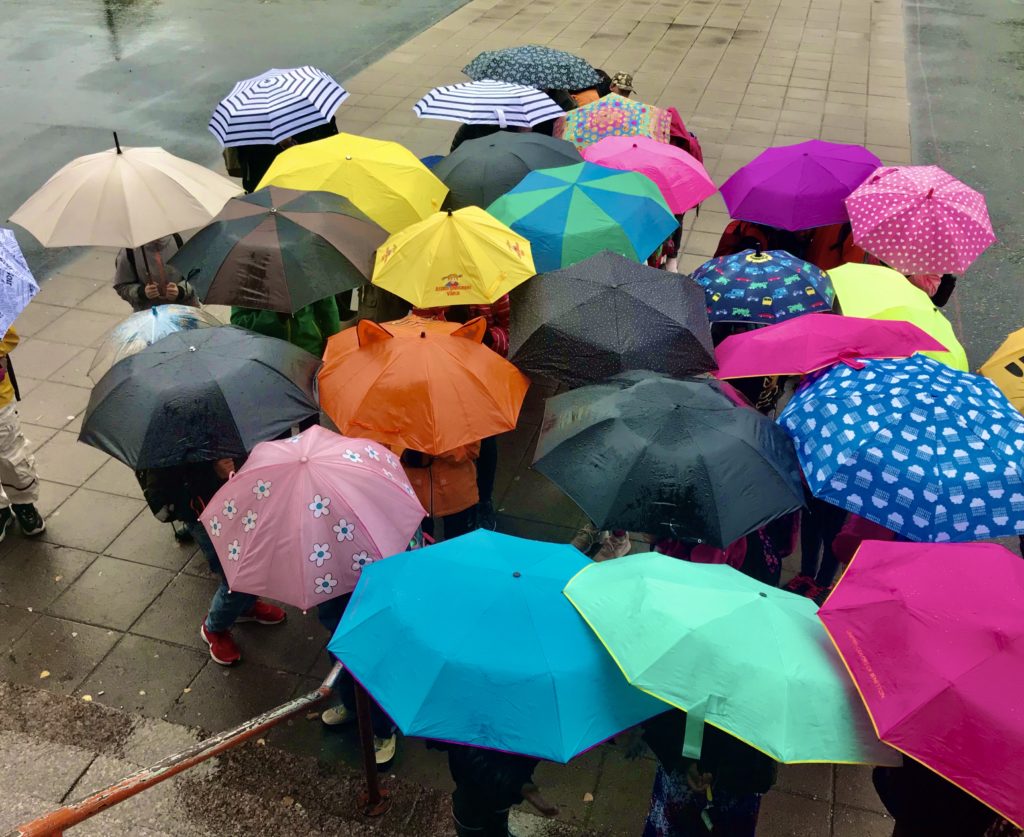 Avattuja sateenvarjoja koulun pihalla rykelmässä, alla lapsia.