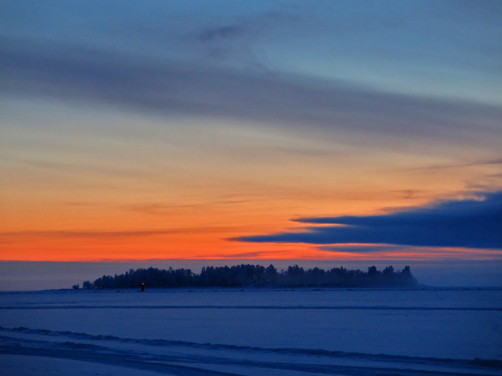 Talvinen kuva, lumi näyttää siniseltä, aurinko punertaa taivasta