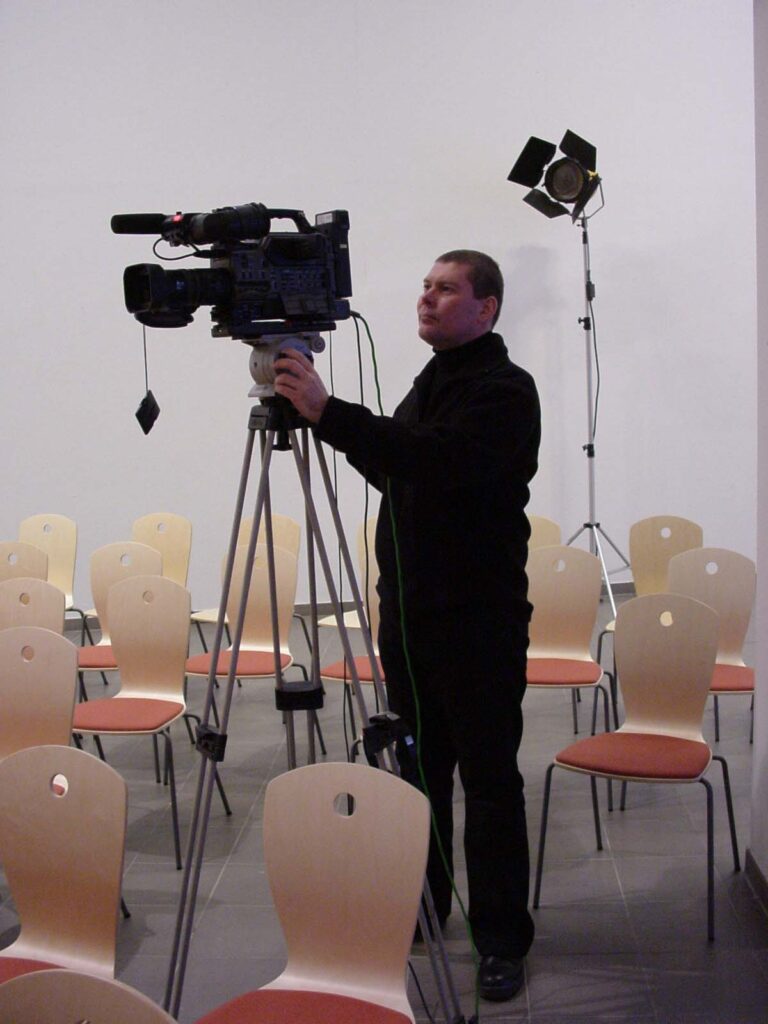Mies TV-kameran takana, ympärillä tyhjiä tuoleja.