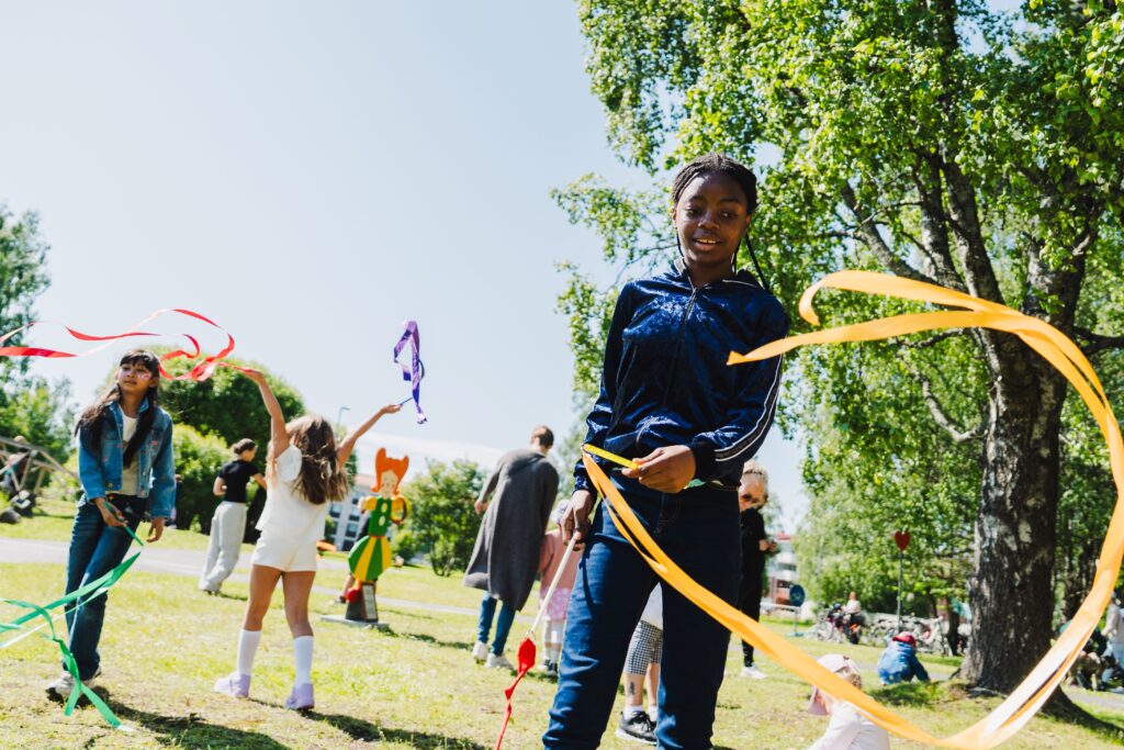 Lapsia kkesäisessä puistossa heiluttamassa värikkäitä nauhoja.