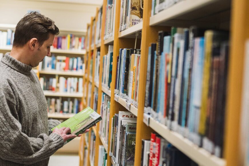 Mies lukee selaa kirjahyllyjen välissä.