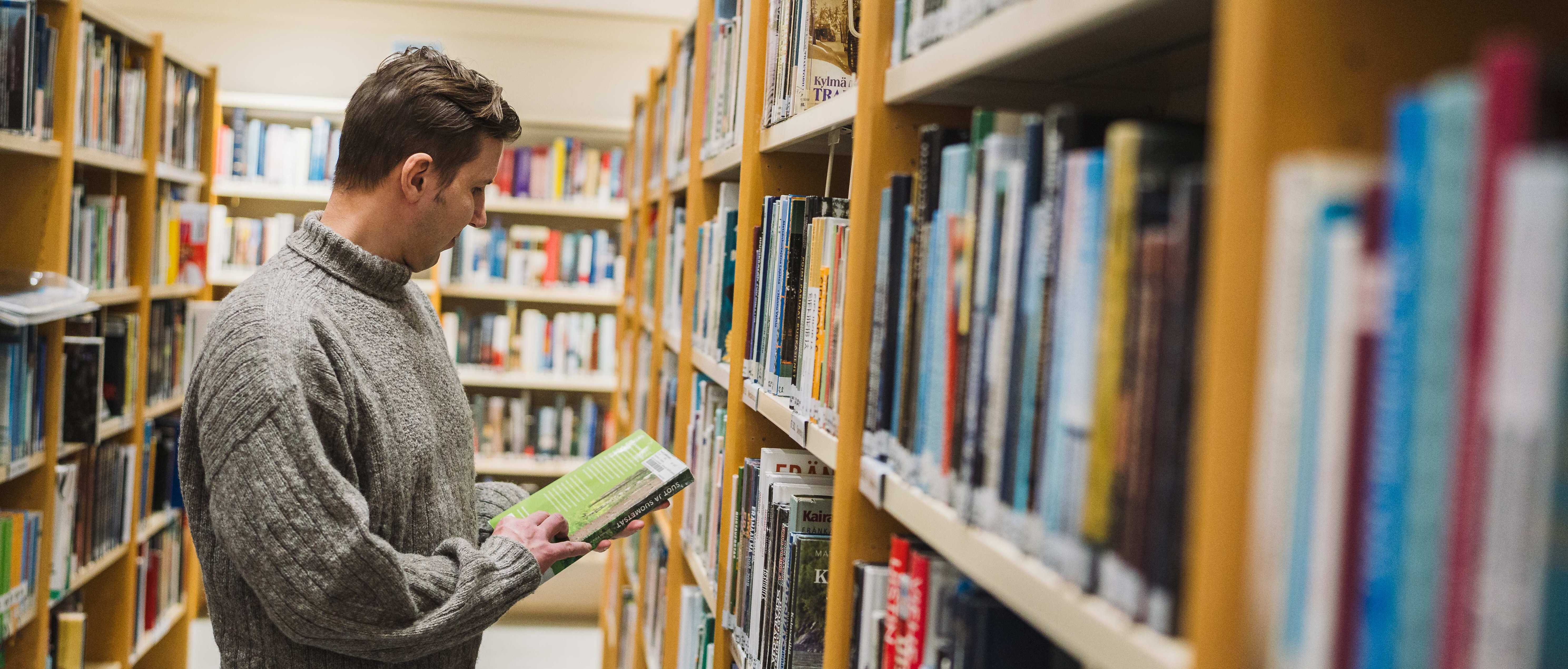 Mies lukee selaa kirjahyllyjen välissä.