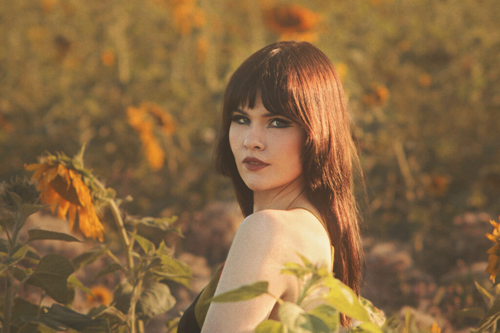 Selin oleva nainen katsoo olan yli kuvaajaan, taustalla auringonkukkapelto.