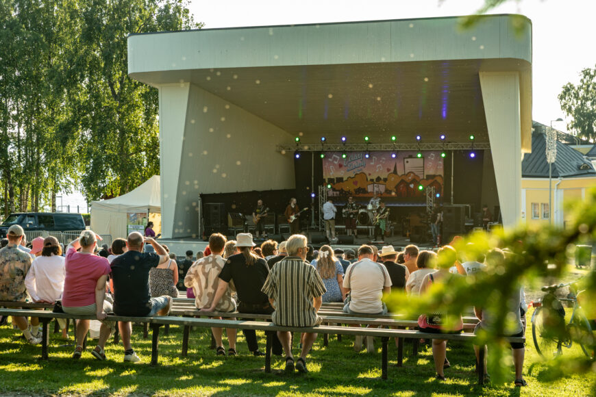 Yleisö istuu penkeillä kesäisellä säällä ulkoilmakatsomossa ison lavan edessä.