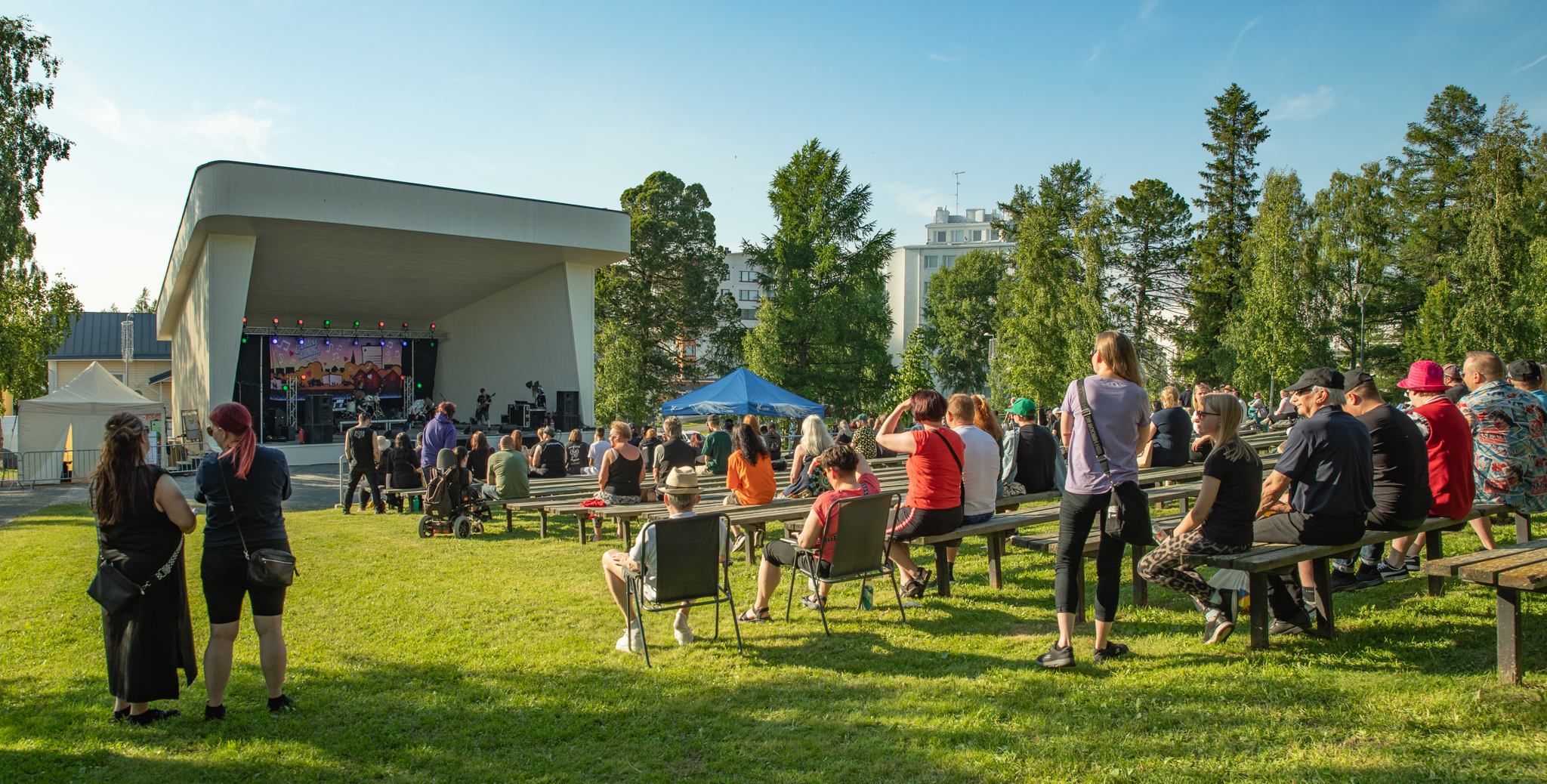 Ihmisiä istumassa ulkoilmakatsomossa Funkkistyylisen lavan edessä, puistomaisemassa.
